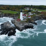 the shore with a lighthouse on it, trees, and town landscape behind