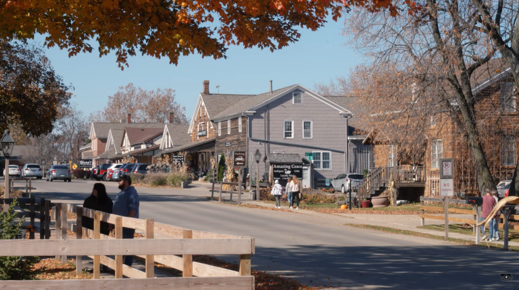 street in a town, buildings on the right side, and people walking on street, other standing behind the tree