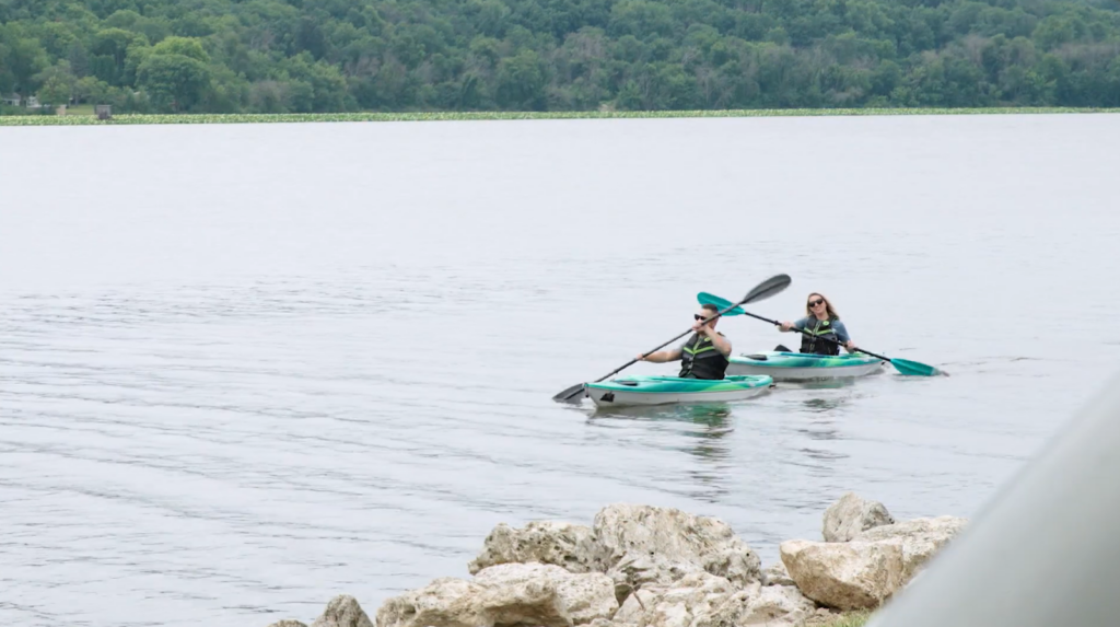 a woman and man on the kayaks with paddles on the lake, the shore with trees behind