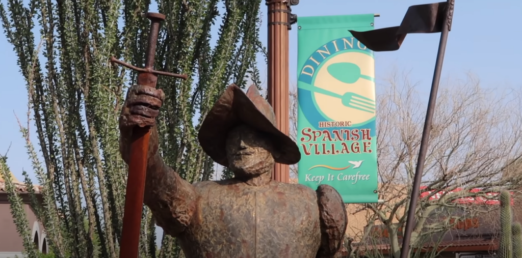 Statue with hat and sword outdoors, surrounded by trees and a banner